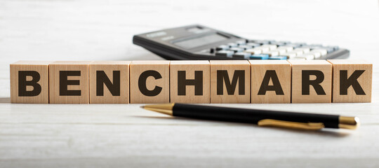 The word BENCHMARK is written on wooden cubes near a calculator and a pen on a light background. Business concept
