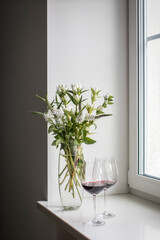 A bouquet of white veronica with white bells in a tall narrow vase on a grey table on the white background. red wine in tall glass glasses.