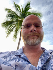 Bald man with beard and glasses under tropical island palm tree smiling and looking down