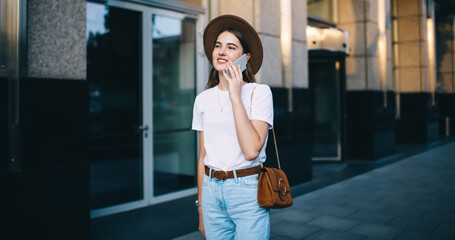 Trendy dressed hipster girl in hat enjoying outdoors walking on city street talking on mobile phone, beautiful 20s woman in stylish apparel satisfied with international roaming connection making call