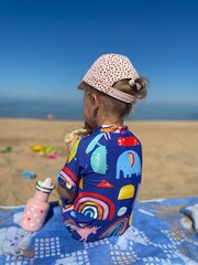 little girl in colorful swimsuit sitting on the blue blanket at the beach on a dummy summer warm day