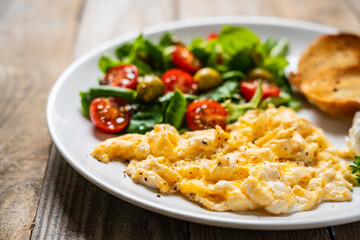 Delicious breakfast - scrambled eggs with fried sausages, cottage cheese and vegetables served on black plate on wooden table
