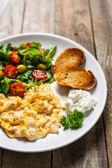 Delicious breakfast - scrambled eggs with fried sausages, cottage cheese and vegetables served on black plate on wooden table
