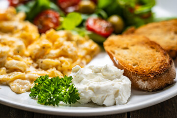 Delicious breakfast - scrambled eggs with fried sausages, cottage cheese and vegetables served on black plate on wooden table
