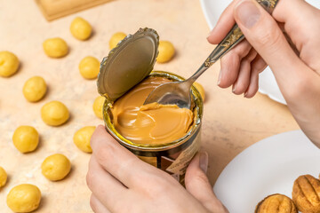 woman's hand takes a teaspoon of boiled condensed milk from a tin can, next to raw cookie dough and ready-made halves for cookie Nuts