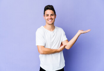 Young caucasian man isolated on purple background showing a copy space on a palm and holding another hand on waist.