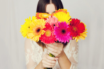 Beautiful woman in white dress hid her face behind large bouquet of colorful bright gerberas.