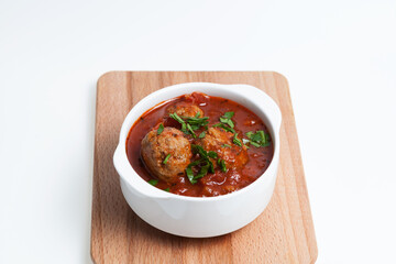 Beef meatballs in white bowl with tomato sauce served with fresh chopped spinach or basil on wooden board. Light background, copy space