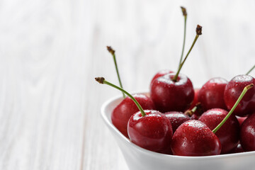 Fresh cherry berries with drops of water in a white bowl on a white wooden table. Close-up macro shot.