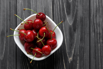 Fresh cherry berries with drops of water in a white bowl on a black wooden table. Close-up macro shot top view.