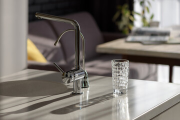 Glass of pure filtered water on the kitchen table in the rays of sunlight. Kitchen faucet and reverse osmosis faucet for water purification. Healthy lifestyle concept. Сlose-up shot, selective focus.