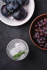 Fresh ice cold carbonated water in glass with rosemary leaf near to wooden bowls with grape and plum fruits, dark stone background, top view