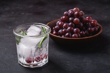 Fresh ice cold carbonated water in glass with rosemary leaf near to wooden bowl with grape berries, dark stone background, angle view