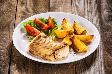 Fish dish - fried cod fillet with potatoes and vegetable salad on wooden table