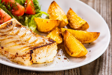 Fish dish - fried cod fillet with potatoes and vegetable salad on wooden table