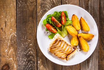 Fish dish - fried cod fillet with potatoes and vegetable salad on wooden table