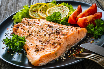 Fried salmon fillet with lettuce, lemon and tomatoes served on black plate on wooden table
