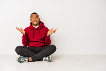 Young latin man sitting on the floor isolated confused and doubtful shrugging shoulders to hold a copy space.