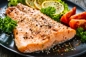 Fried salmon fillet with lettuce, lemon and tomatoes served on black plate on wooden table
