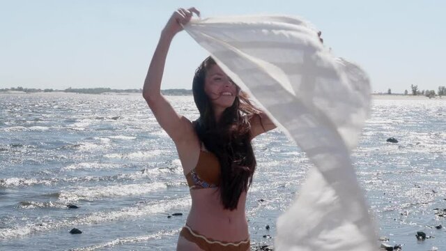 Swarthy woman in swimsuit with long hair plays with scarf fluttering in wind on beach