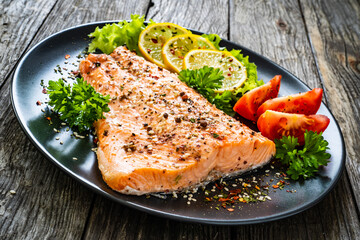 Fried salmon fillet with lettuce, lemon and tomatoes served on black plate on wooden table
