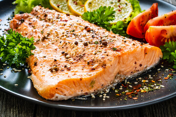 Fried salmon fillet with lettuce, lemon and tomatoes served on black plate on wooden table
