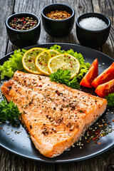 Fried salmon fillet with lettuce, lemon and tomatoes served on black plate on wooden table
