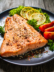 Fried salmon fillet with lettuce, lemon and tomatoes served on black plate on wooden table
