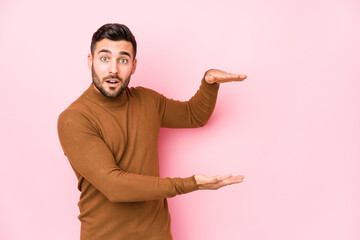 Young caucasian man against a pink background isolated shocked and amazed holding a copy space between hands.