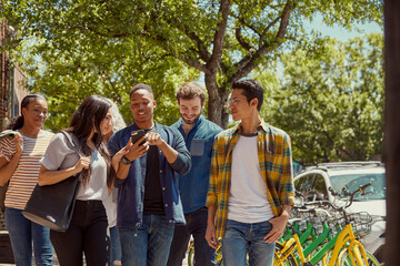 Group of young co-workers walking down sidewalk together, young man showing friends something on cell phone 