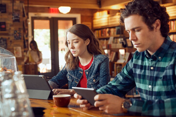 Young man and woman sitting at counter in cafe bookstore using digital tablets 