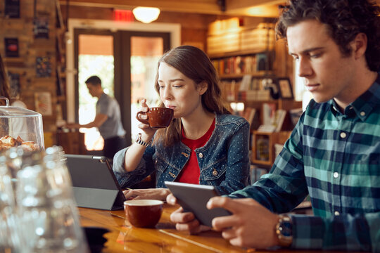 Young man and woman sitting at counter in cafe bookstore using digital tablets , woman having sip of coffee  