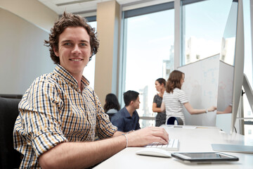 Young man using computer in office looking towards camera, co-workers having discussion in background 