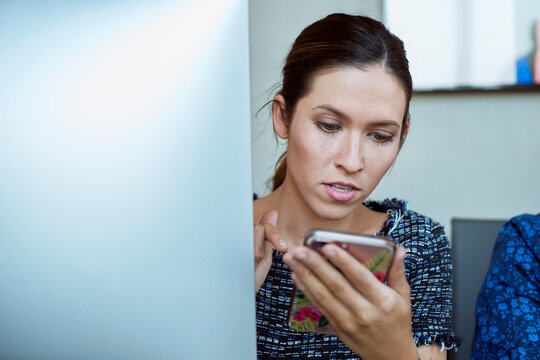 Young woman checking smart phone , while sitting at computer in office