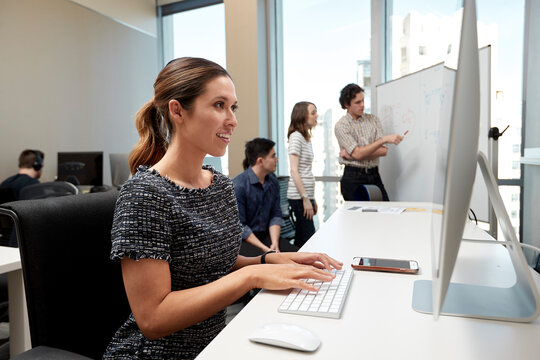 Young woman using computer in office typing on keyboard, co-workers having discussion around dry erase board in background 