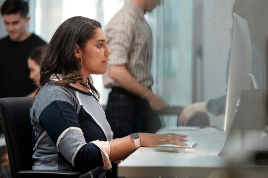 Young woman using computer in office typing on keyboard, co-workers having discussion around dry erase board in background 