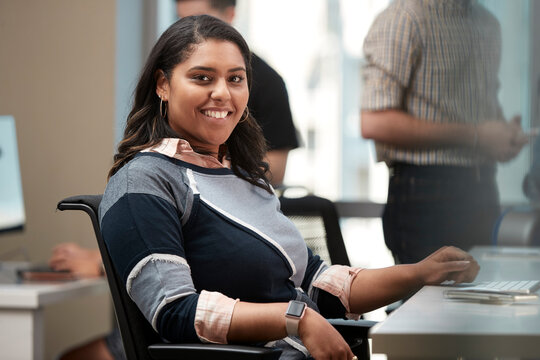 Young woman sitting at computer in office looking to the camera smiling, co-workers having discussion around dry erase board in background 