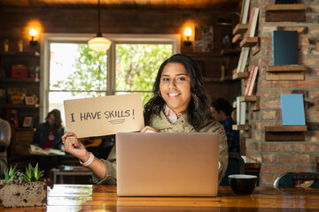 Portrait of young woman sitting at table in coffee shop bookstore with laptop computer , holding hand made sign that reads I have skills