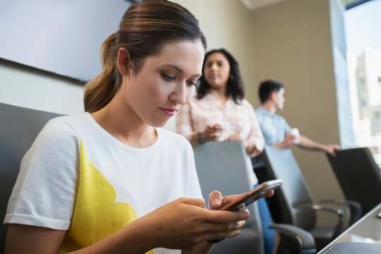 Young woman in conference room checking mobile phone, with co-workers in background 