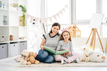 Beautiful mother and daughter reading book sitting on floor in kids room