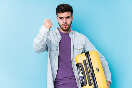 Young caucasian traveler man holding a suitcase isolated showing fist to camera, aggressive facial expression.