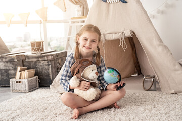 Happy little girl with globe and teddy bear sitting on floor in front of wigwam © Ievgen Skrypko