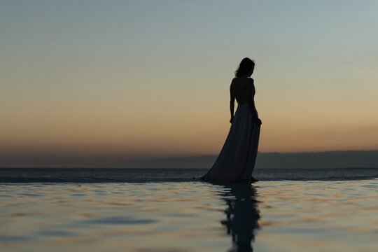 Slim woman silhouette walking on the pool edge against sunset sky