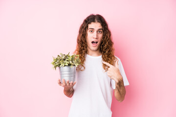 Long hair caucasian man holding a plant surprised pointing at himself, smiling broadly.