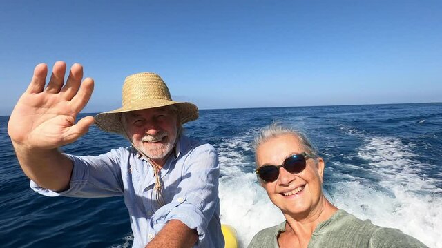 couple of cute mature people or senior enjoying and having fun together in the middle of the sea or ocean with a small boat or dinghy - woman pensioner taking a selfie with her old man driving the boa