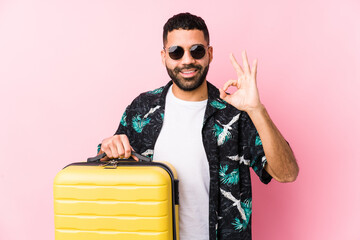Young latin man holding a suitcase cheerful and confident showing ok gesture.