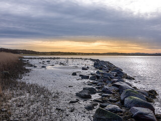View of a coastline with the early morning sun in the background. A row of boulders follow a perspective line into the photo and it is a cloudy but calm morning. It is shot in early spring.