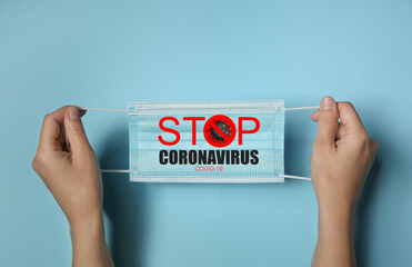 Woman holding medical mask with text Stop Coronavirus on light blue background, closeup. Protective...
