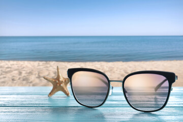 Fototapeta na wymiar Starfish and stylish sunglasses on light blue wooden table near sea with sandy beach