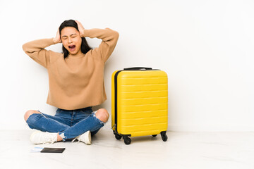 Young chinese traveler woman sittting on the floor with a suitcase isolated covering ears with hands trying not to hear too loud sound.